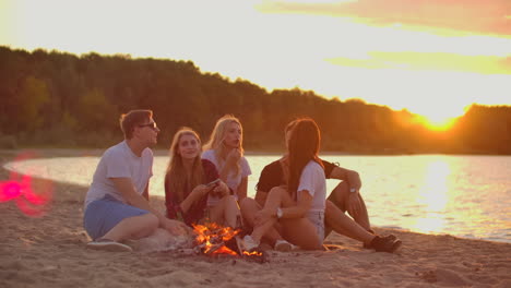 The-students-are-sitting-around-bonfire-on-the-beach.-They-are-talking-to-each-other-and-clink-beer-at-sunset-and-enjoying-the-summer-evening-on-the-river-coast.